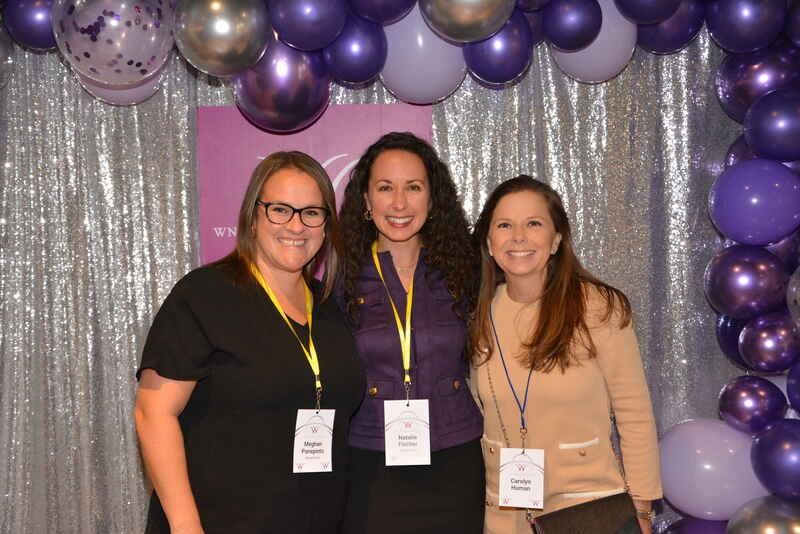 Three women are posing for a picture in front of purple balloons.