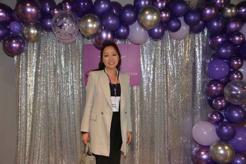 A woman is standing in front of a wall covered in purple and gold balloons.