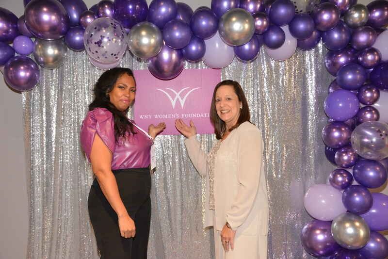 Two women are standing in front of a wall of purple balloons.