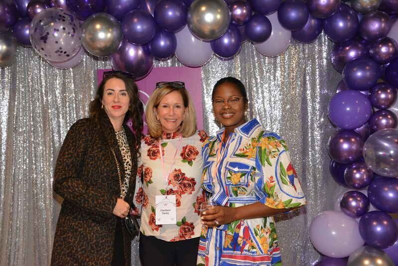 Three women are posing for a picture in front of a purple balloon arch.