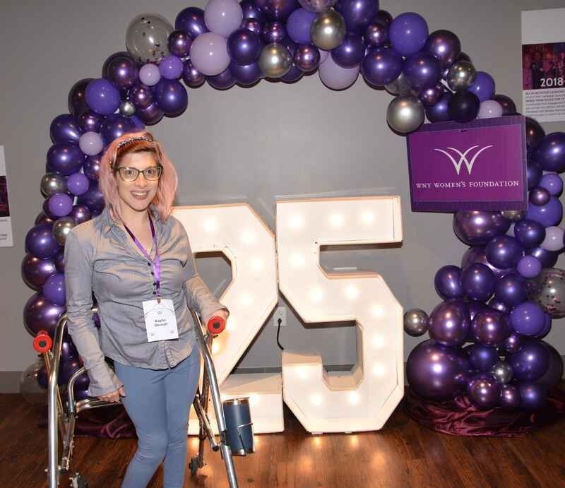 A woman in a wheelchair is standing in front of a purple balloon arch.
