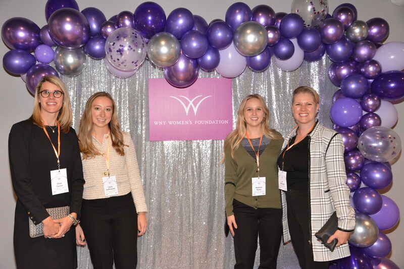 A group of women are posing for a picture in front of a wall of purple balloons.