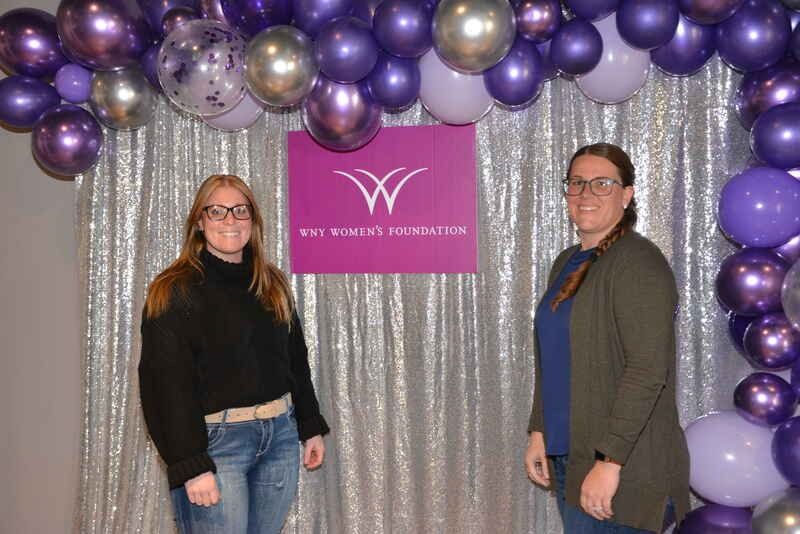Two women are standing in front of a purple balloon arch.