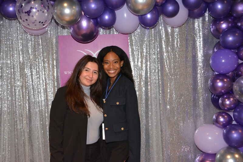 Two women are posing for a picture in front of a purple balloon arch.