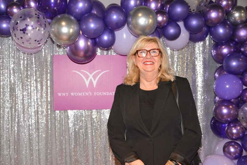 A woman is standing in front of a wall of purple balloons.