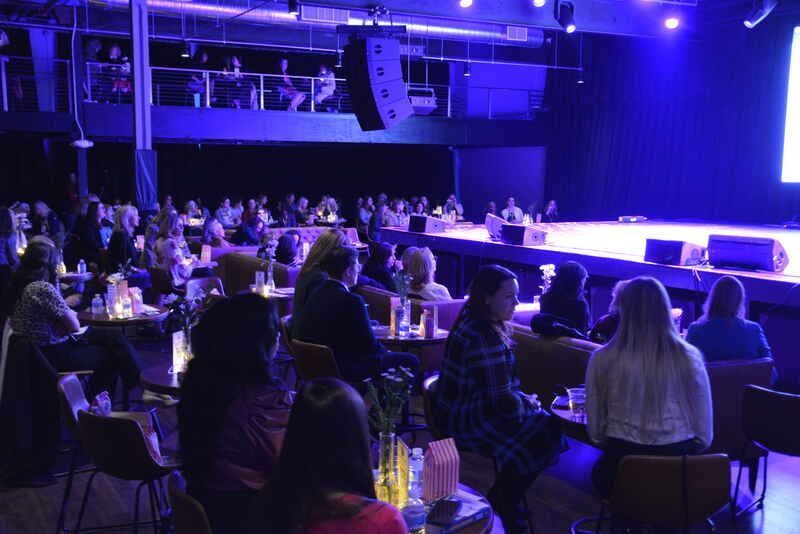 A group of people are sitting at tables in front of a stage.