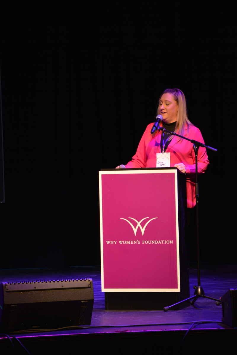 A woman in a red jacket is standing at a podium speaking into a microphone