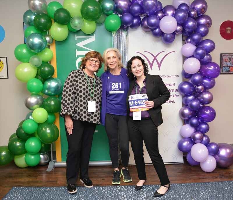 Three women are posing for a picture in front of purple and green balloons