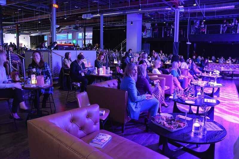 A group of people are sitting at tables in a large room with purple lights.