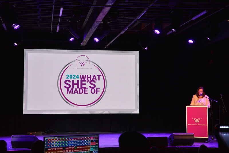 A woman is standing at a podium in front of a large screen that says `` what she 's made of ''.