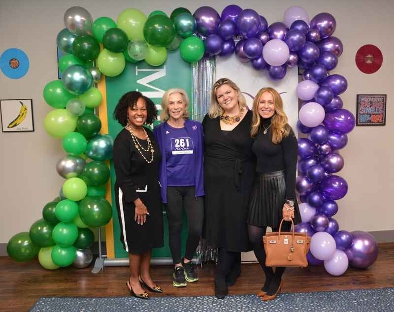 Four women are posing for a picture in front of a green and purple balloon arch.