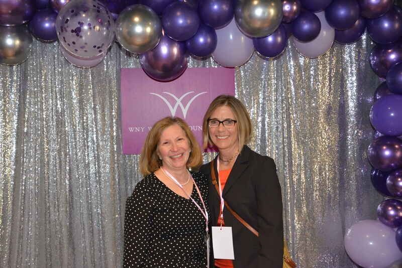Two women are posing for a picture in front of purple balloons.