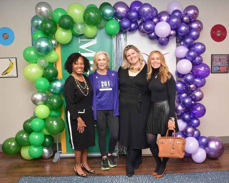 Four women are posing for a picture in front of a green and purple balloon arch.