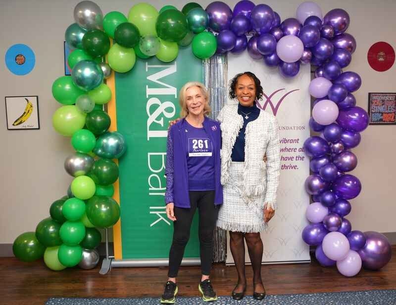 Two women are posing for a picture in front of a green and purple balloon arch.