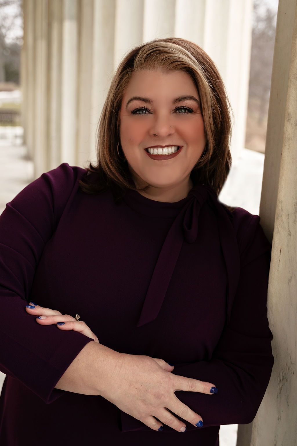 A woman in a purple dress is leaning against a column and smiling.