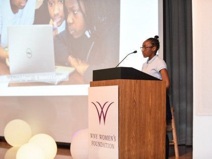 A woman is giving a speech at a podium with a dell laptop on the screen behind her.