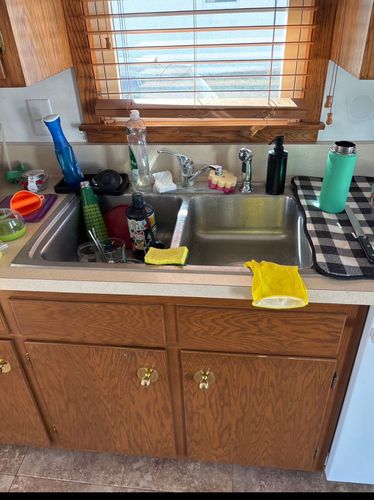 Kitchen sink with dishes, soap, and cleaning supplies under a window with blinds.