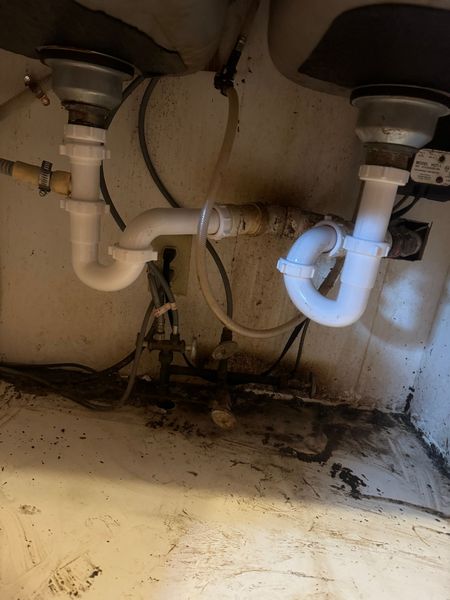 Under-sink plumbing with white pipes and visible water damage, stains, and debris on the cabinet floor