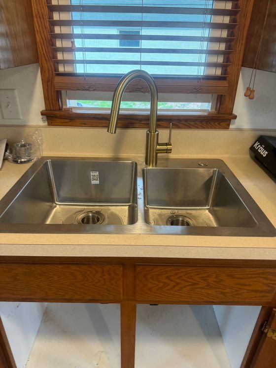 Stainless steel double sink under a wood-framed window with a brass faucet in a kitchen countertop.