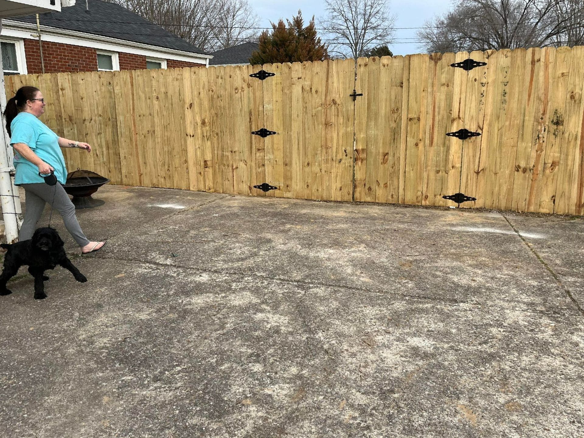 Woman walking black dog on leash in concrete yard, wooden fence by Allen's Tree and Fence.