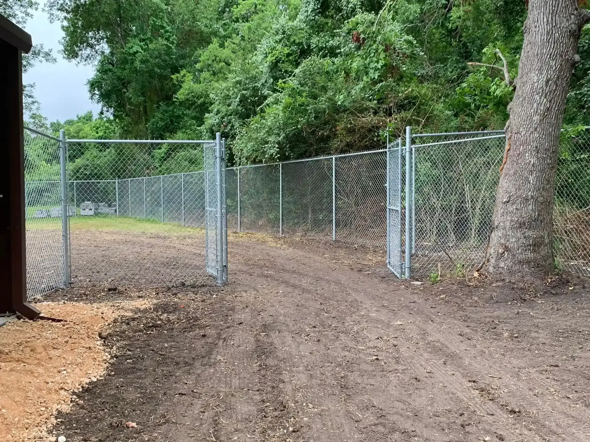 Open chain-link gate in a dirt clearing, bordered by a fence, by Allen's Tree and Fence.