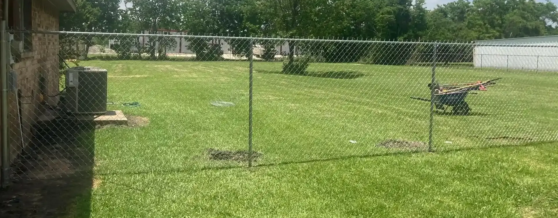A person with a wheelbarrow in a grassy yard, enclosed by a chain link fence.