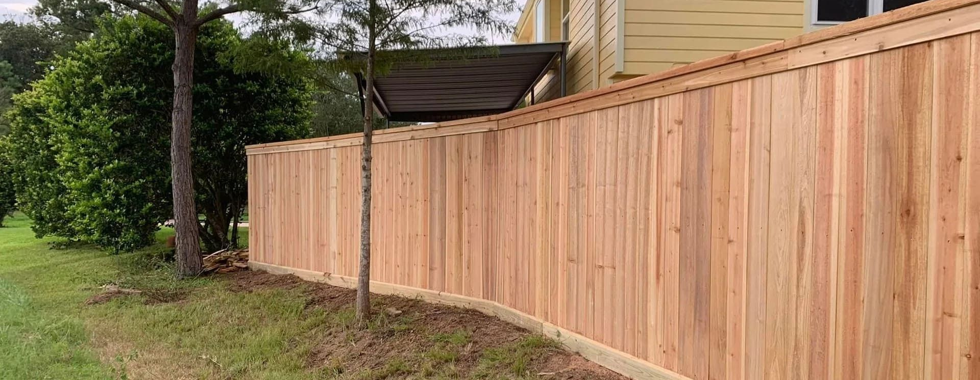 Wooden fence alongside a grassy yard, trees, and a yellow house.