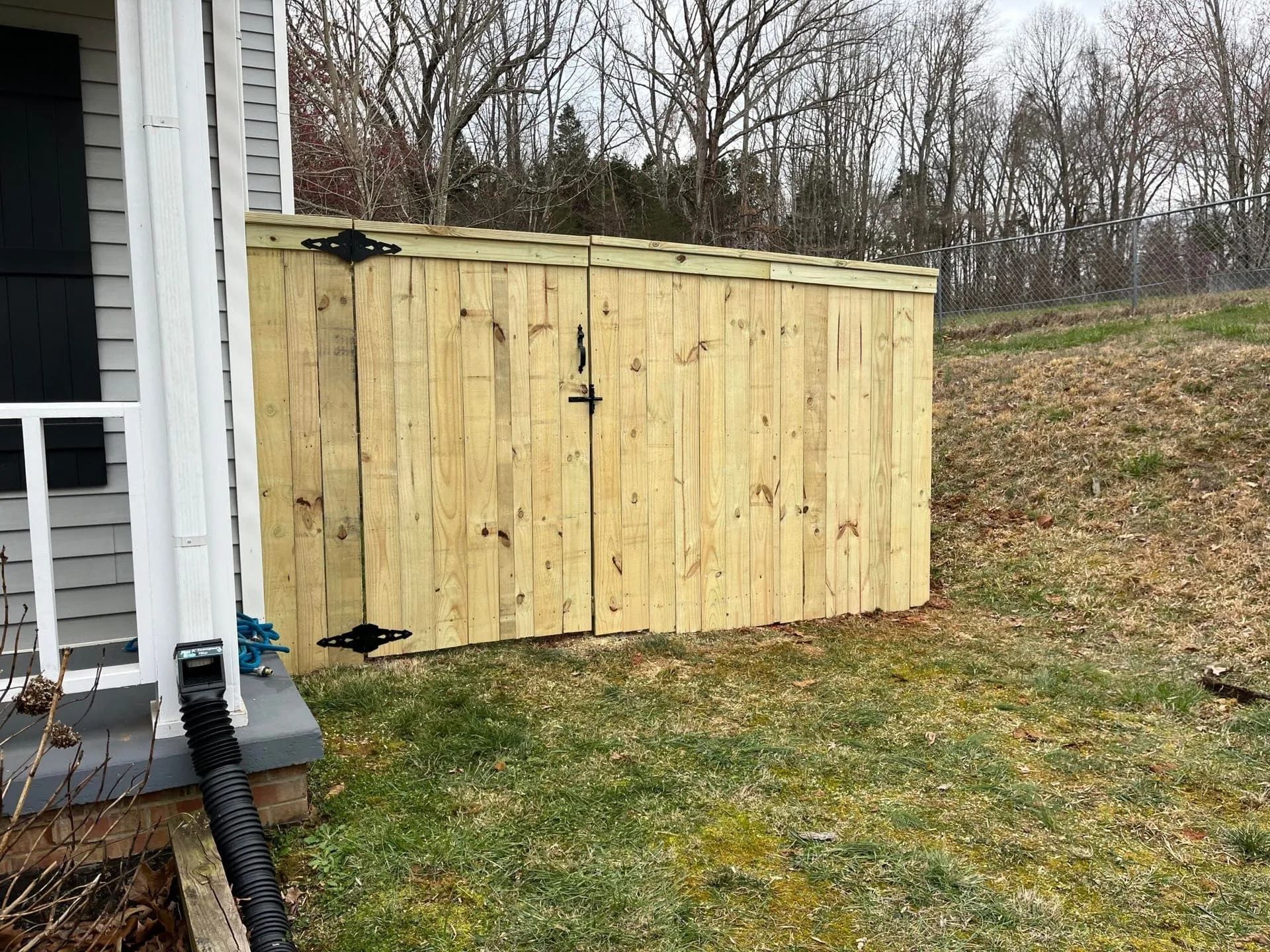 Wooden trash enclosure next to a house with a black downspout; grassy hill in the background by Allen's Tree and Fence.