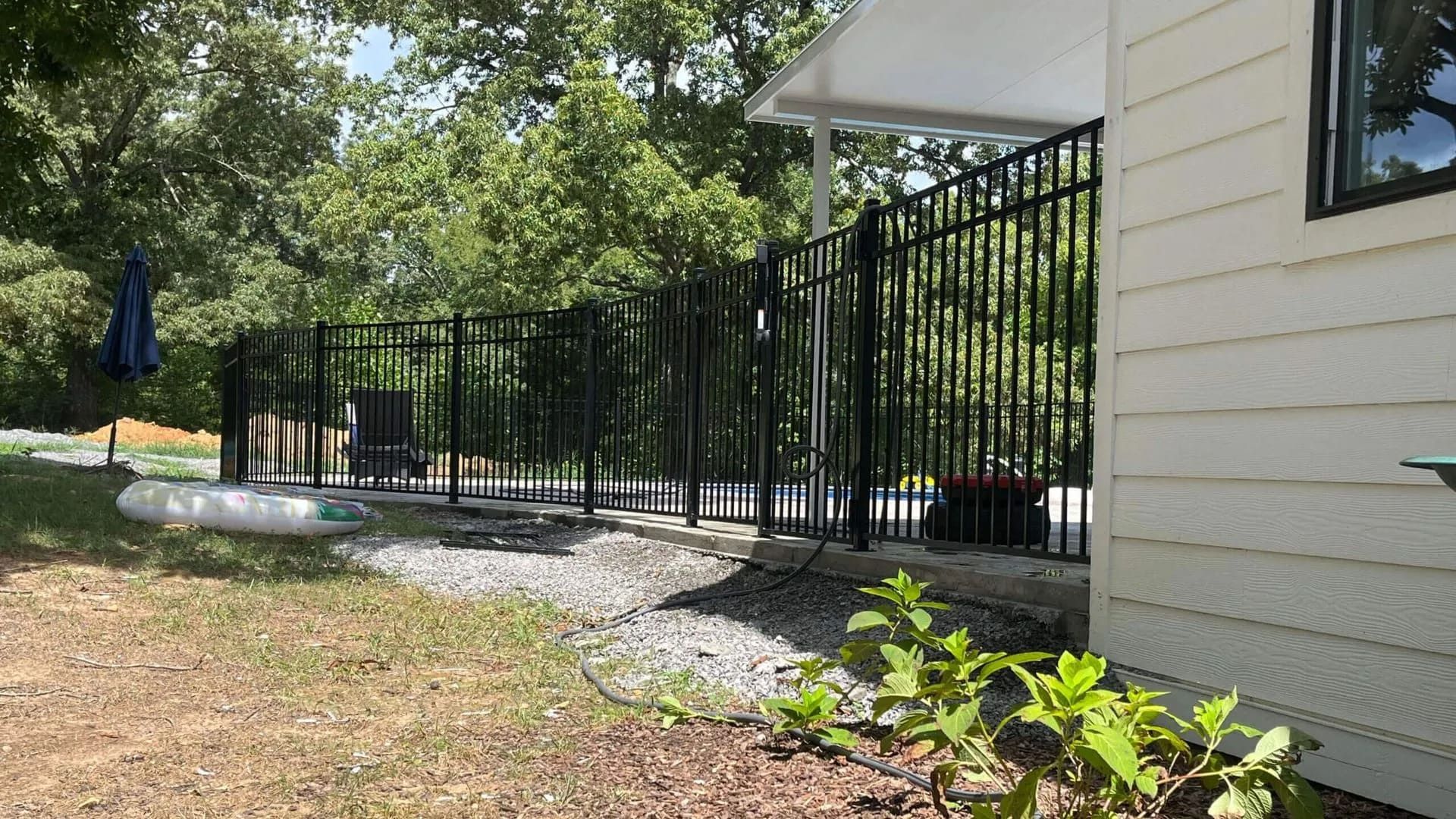 Black fence along a house, gravel bed, and trees in the background on a sunny day.