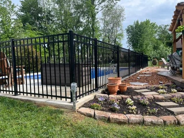 Black metal fence encloses a backyard pool. Stone walkway, flower bed, and lawn surround it.
