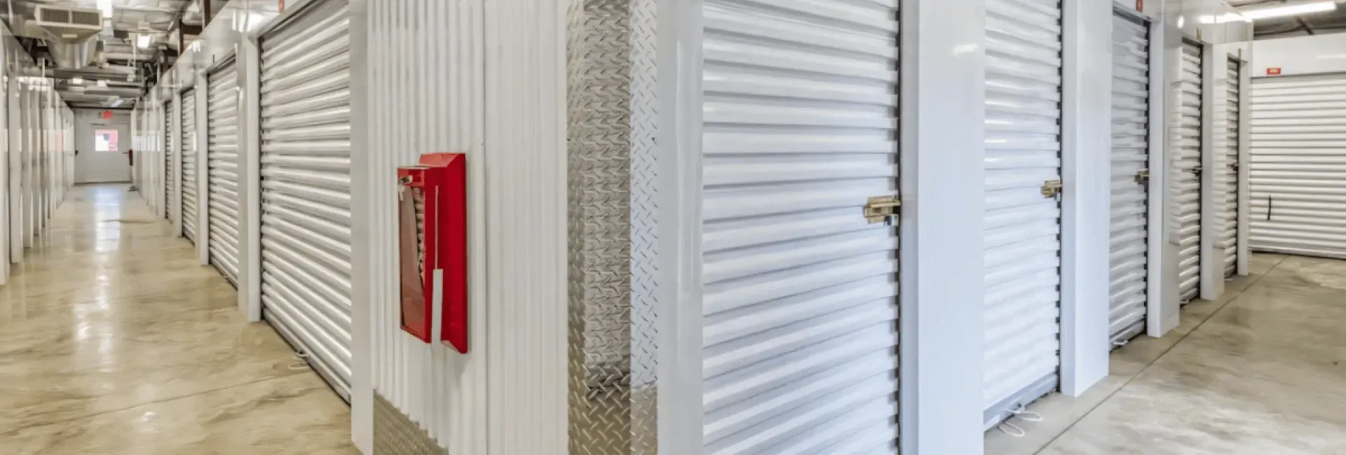 Interior of a storage facility hallway, with white storage units and a red fire alarm.