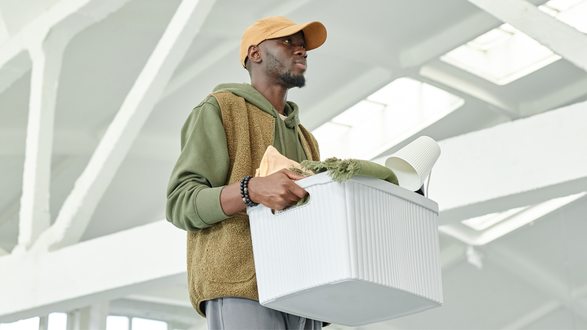 A person in a green hoodie and tan cap walks through a bright building while carrying a plastic bin filled with items.