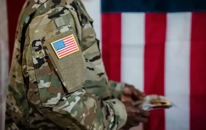 Soldier's arm with US flag patch, holding food, with American flag background.