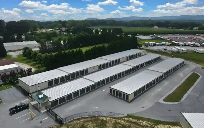 An aerial view of a self-storage facility featuring three rows of long buildings with metal roofs in a gated paved lot.