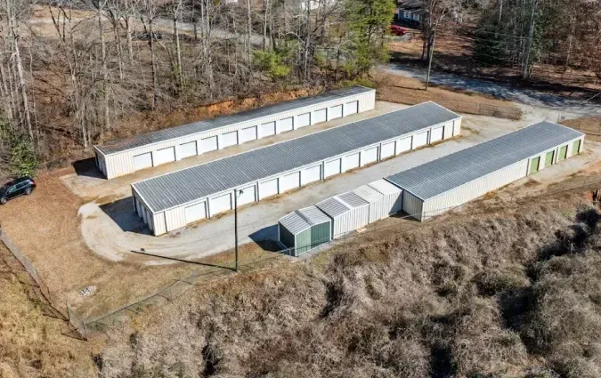 Aerial view of long white storage buildings on a rural dirt lot beside trees and brush.