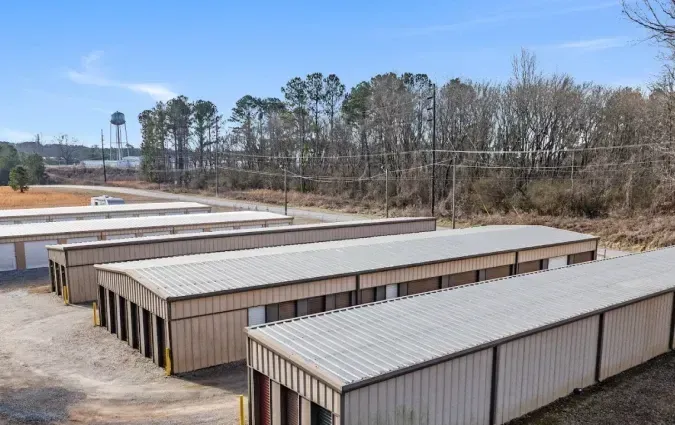 Rows of tan storage units with metal roofs in a gravel lot, with trees and a blue sky behind them