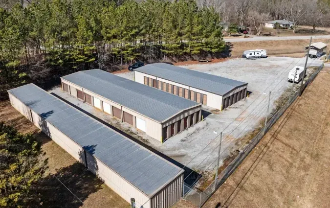 Aerial view of several tan storage units with gray roofs in a paved lot beside trees and fields