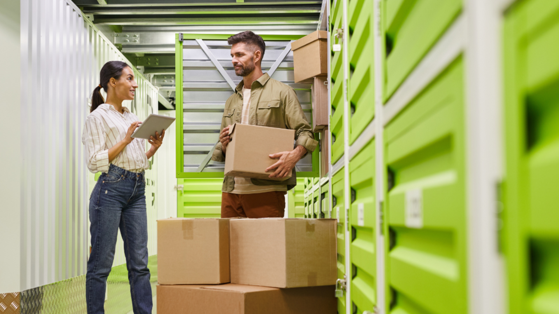 A woman holding a tablet speaks to a man carrying a box in a hallway lined with bright green storage unit doors.