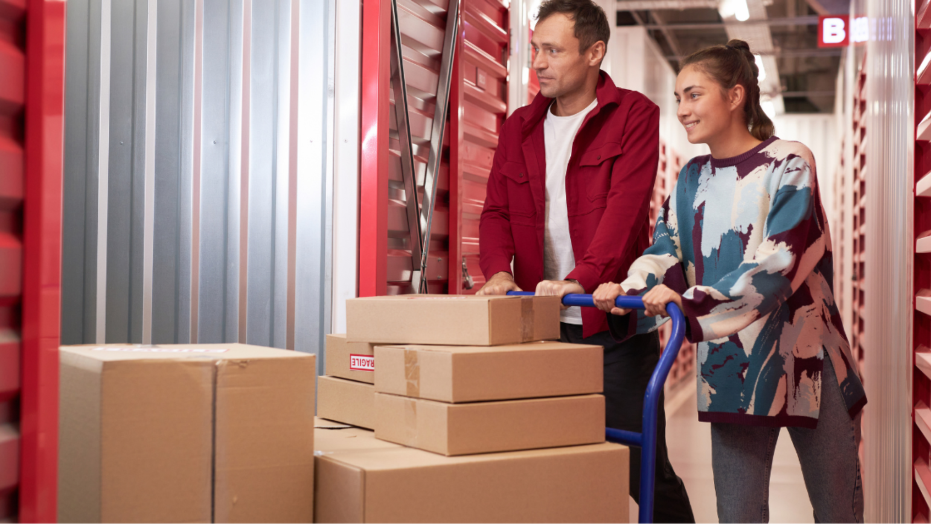 A person in a red jacket and a person in a patterned sweater push a cart full of cardboard boxes down a storage unit hall.