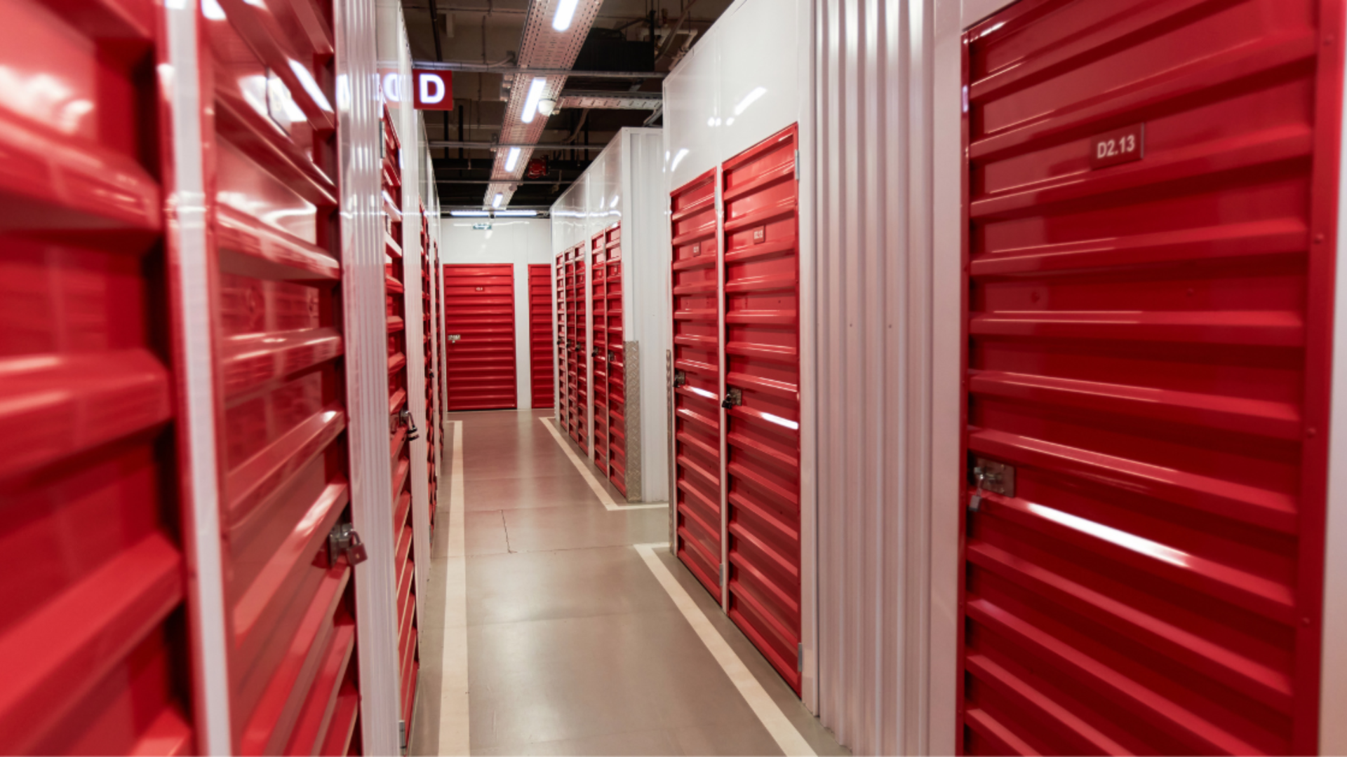 A brightly lit indoor corridor featuring rows of bright red metal self-storage units on both sides.