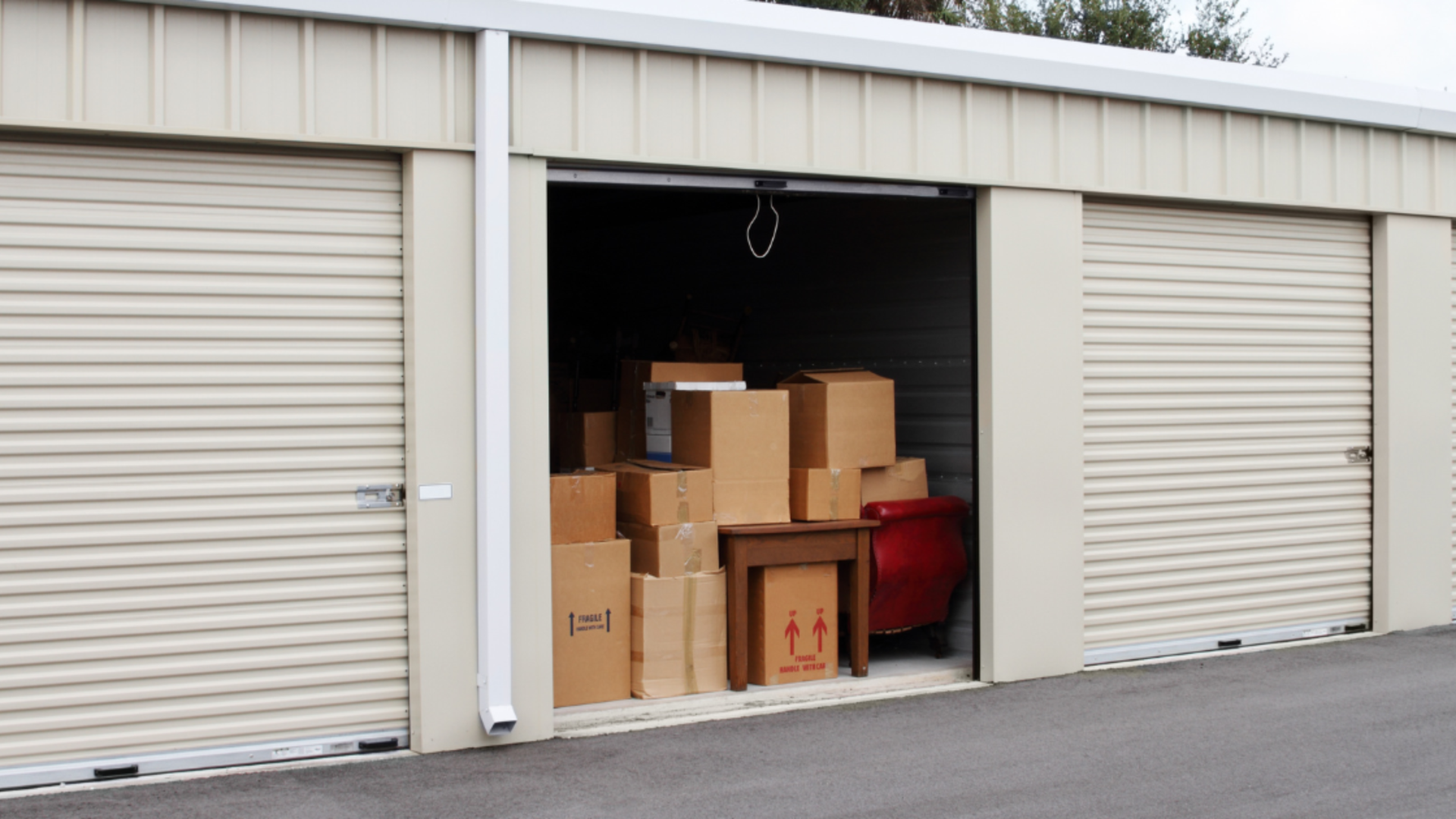 A storage unit with an open roll-up door, filled with stacked cardboard boxes and a small table next to a red armchair.