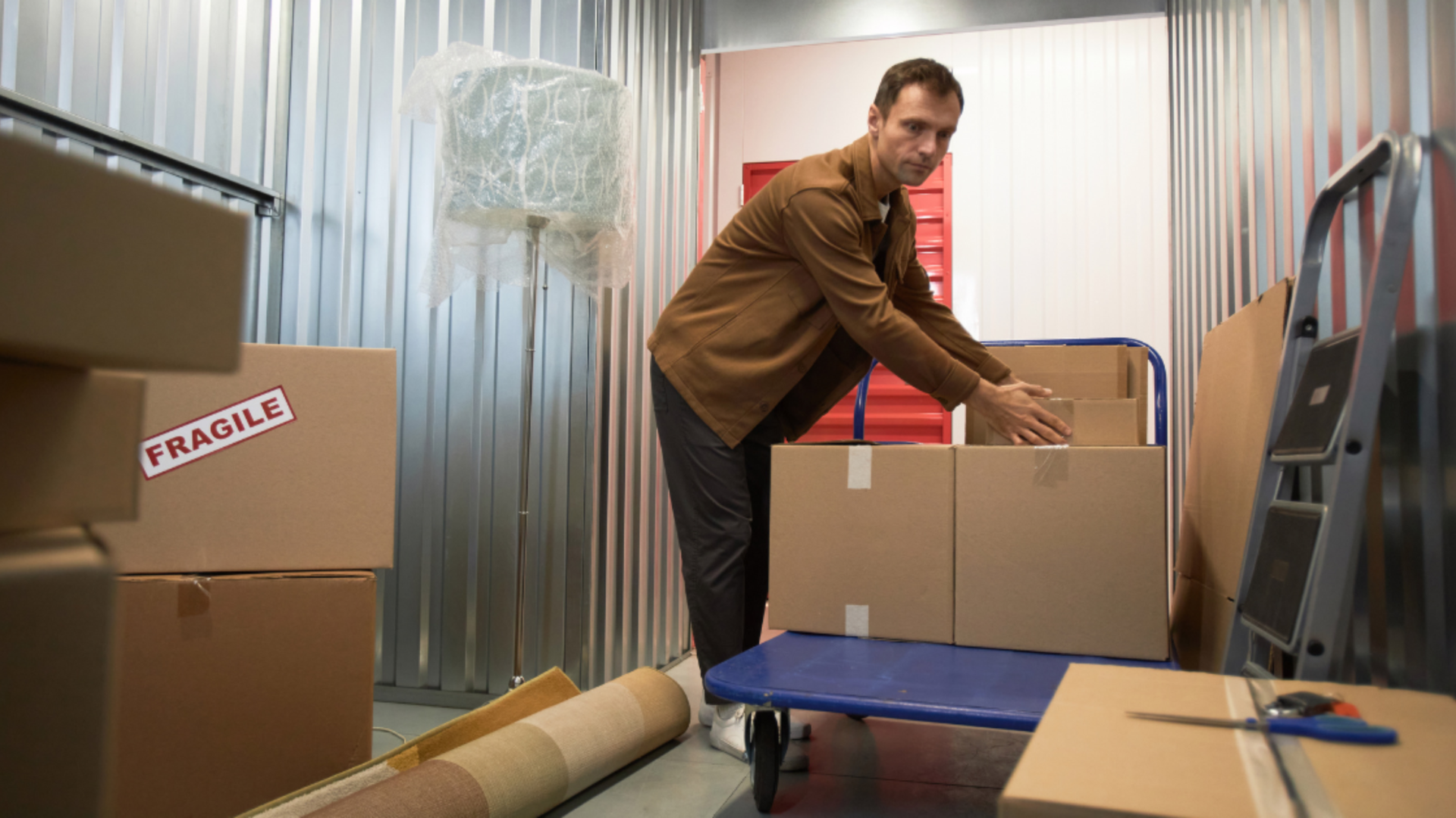 A person in a brown jacket arranges cardboard boxes on a blue cart inside a self-storage unit.