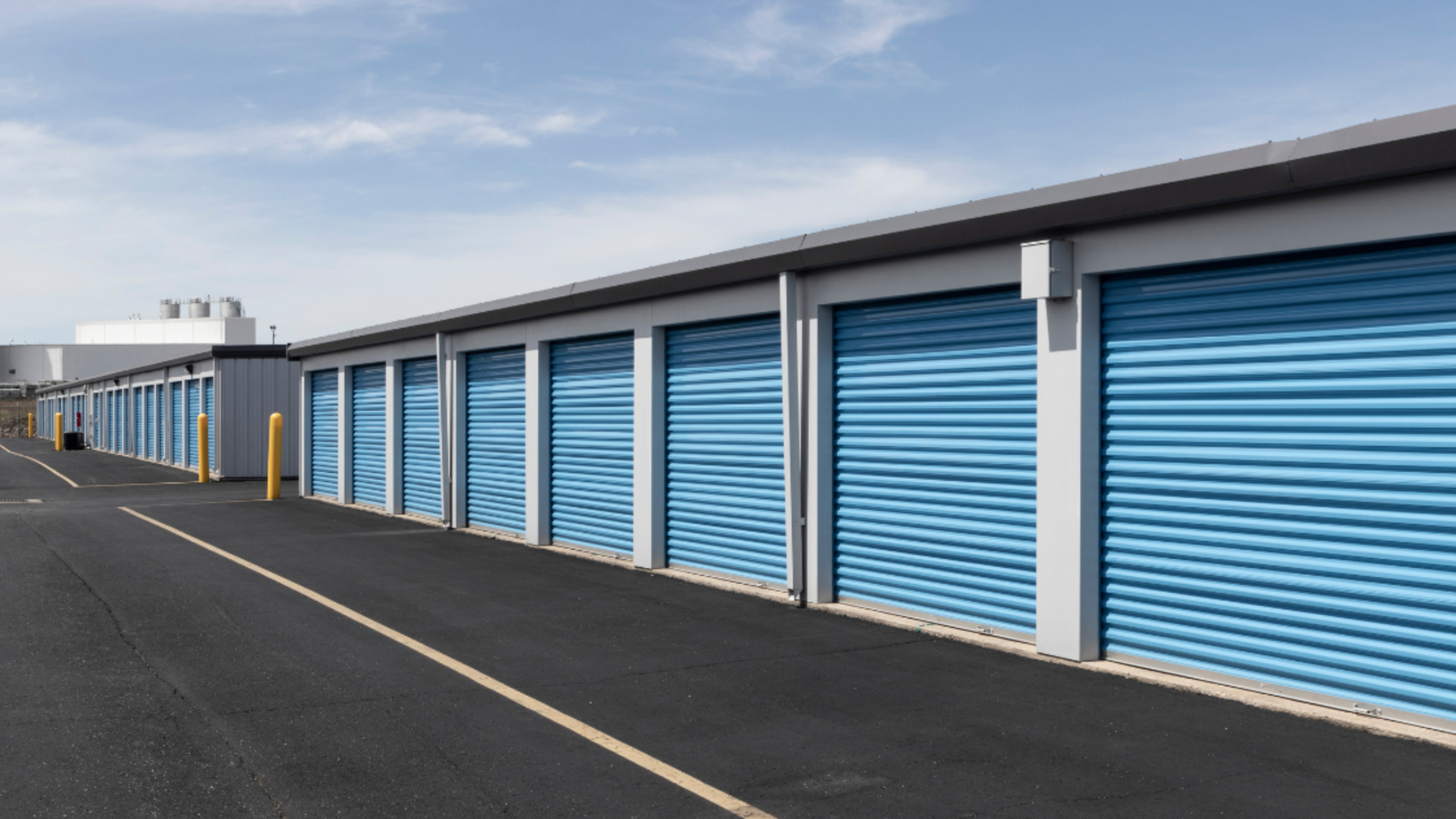 A row of storage units with bright blue roll-up doors along a paved drive under a clear blue sky.
