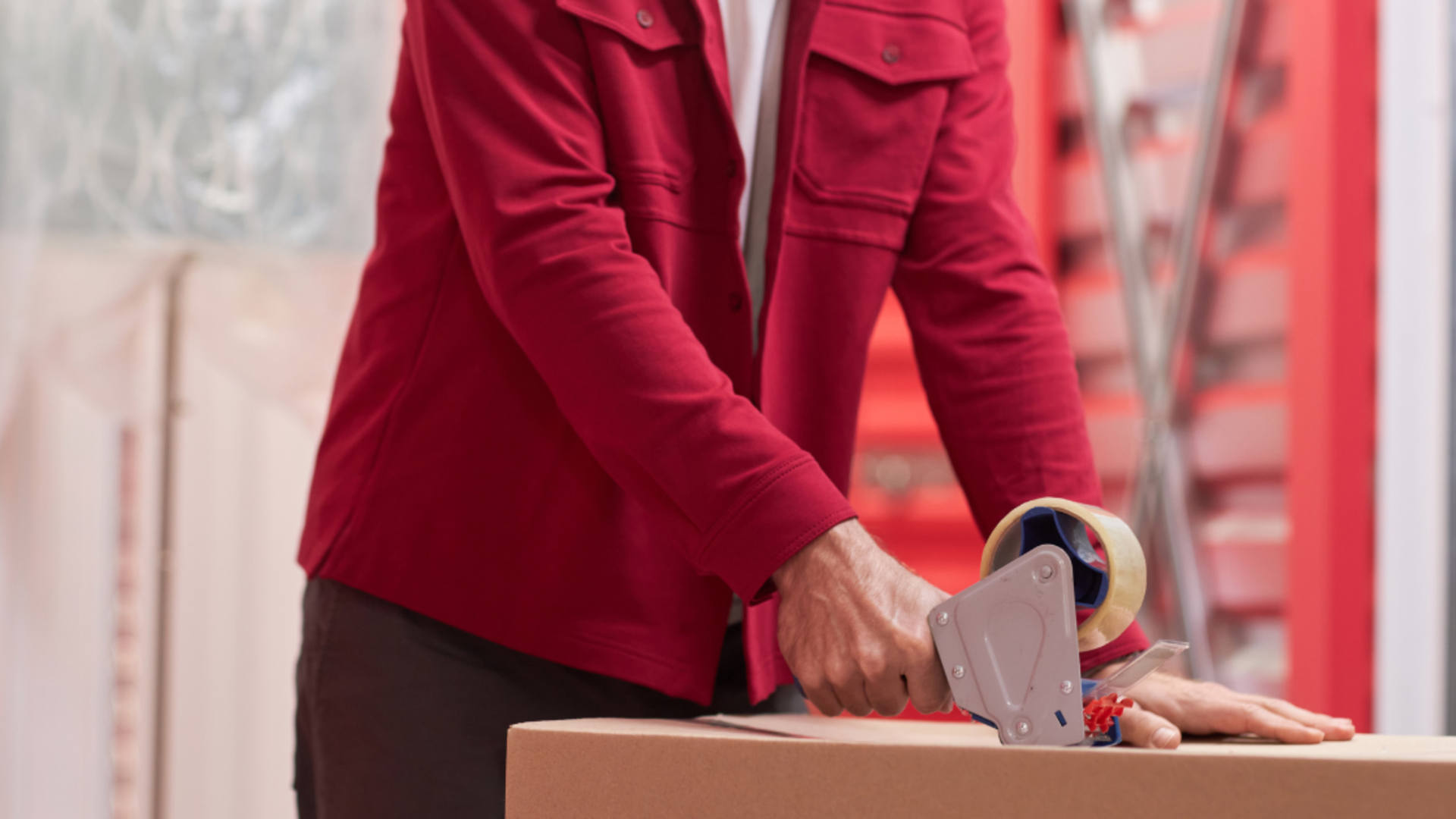 A person wearing a red shirt uses a handheld tape dispenser to seal a cardboard box.