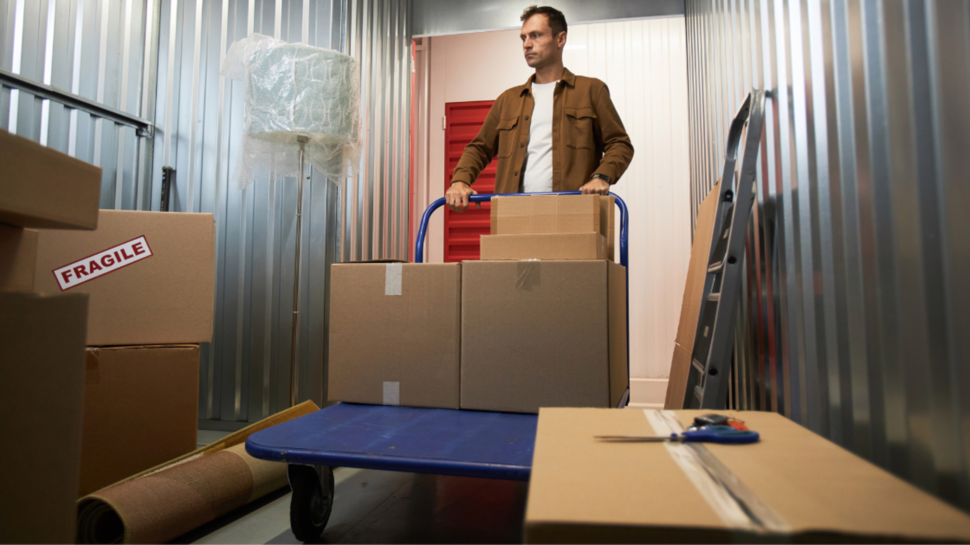 A person in a brown shirt moves boxes on a blue utility cart inside a corrugated metal storage unit.