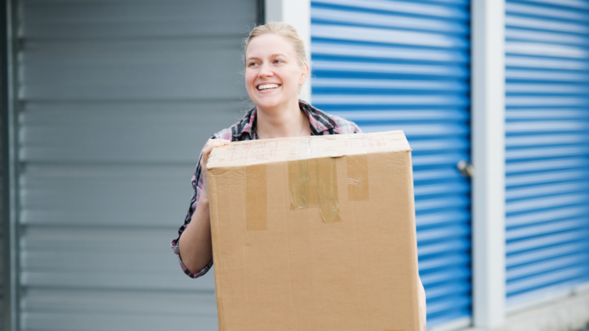 Smiling person carrying a cardboard box in front of blue storage unit doors.
