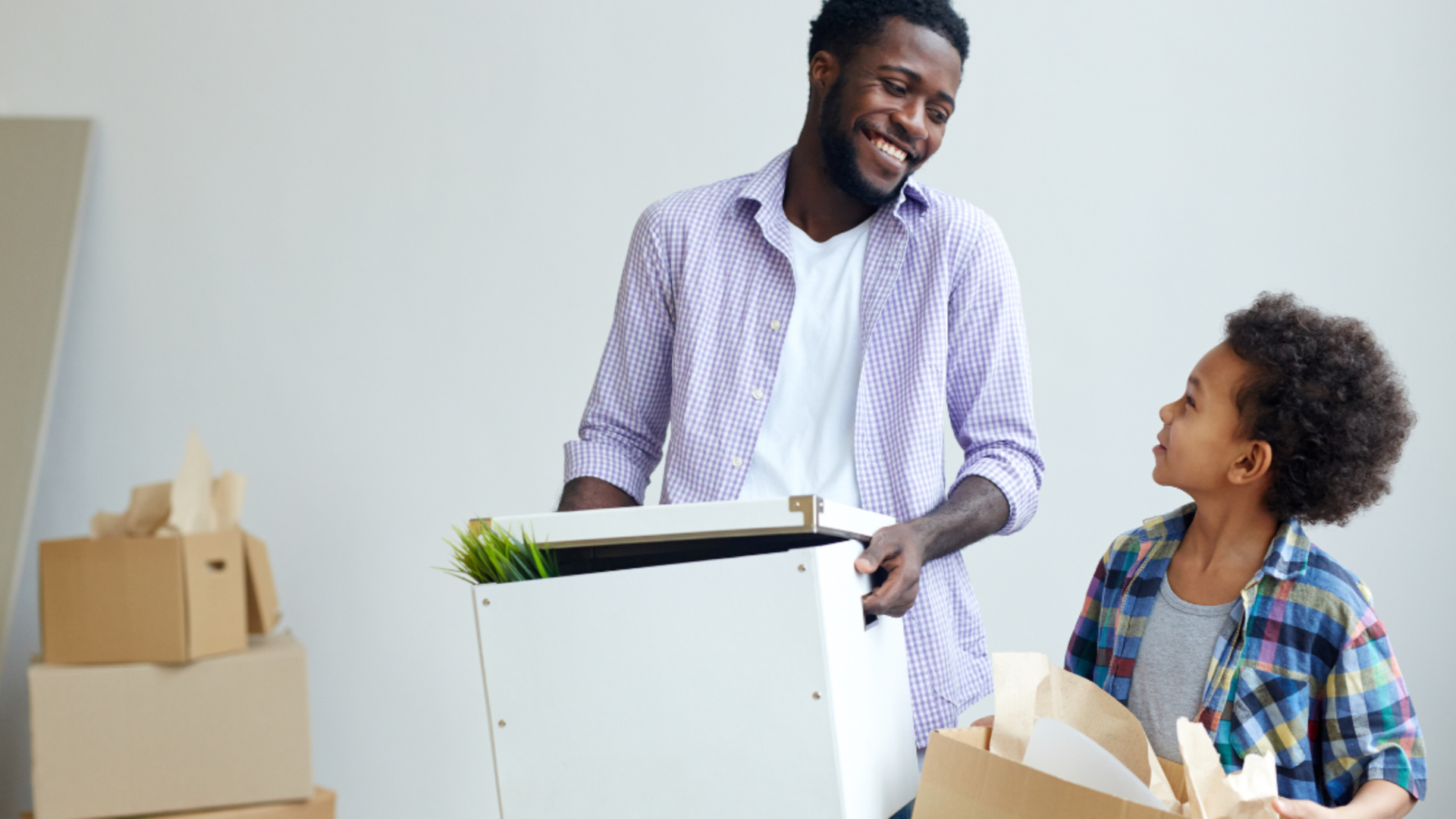 A smiling adult and child holding cardboard boxes while moving into a new home.