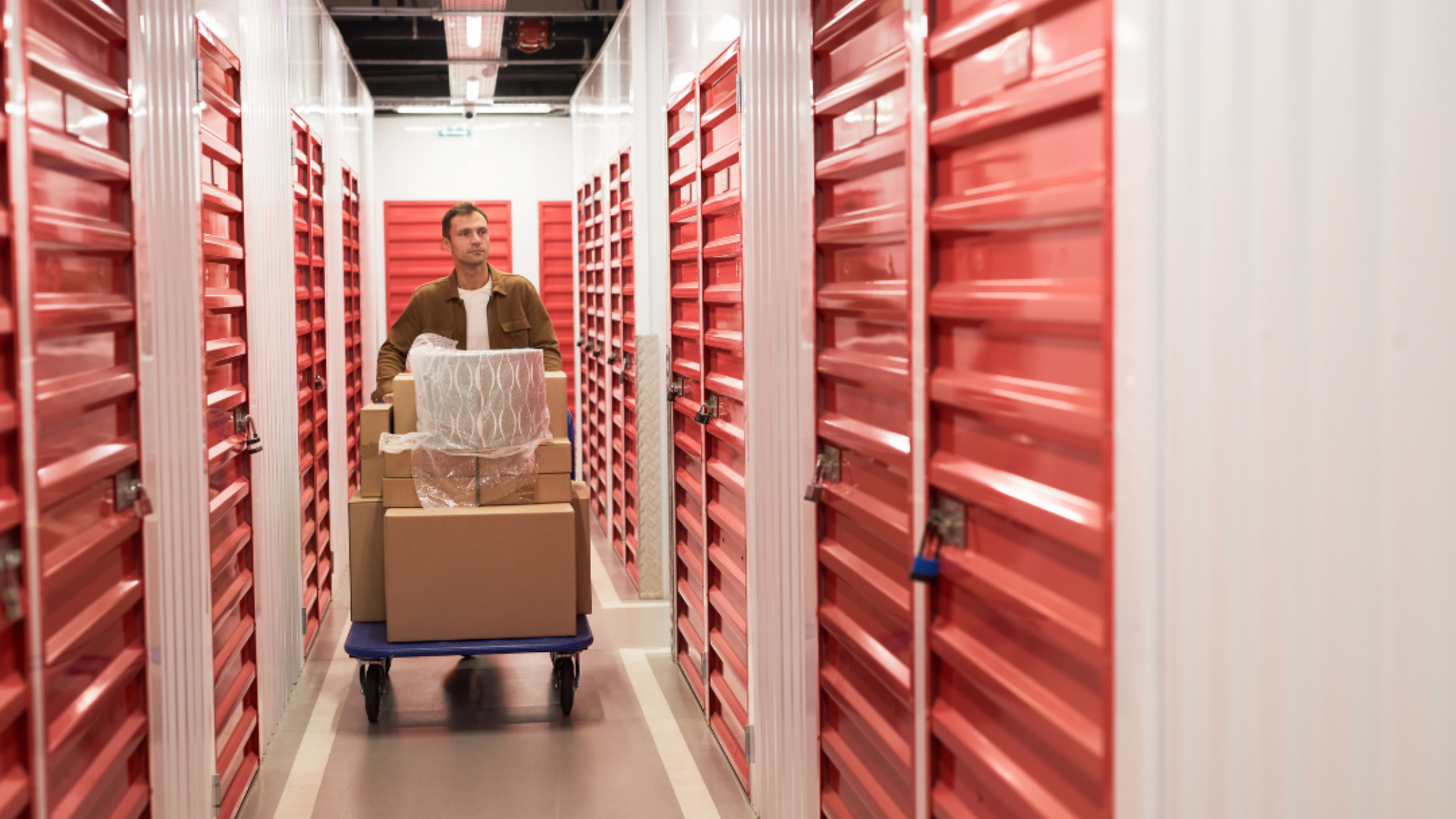 A person pushes a cart loaded with cardboard boxes through a narrow hallway lined with red self-storage unit doors.