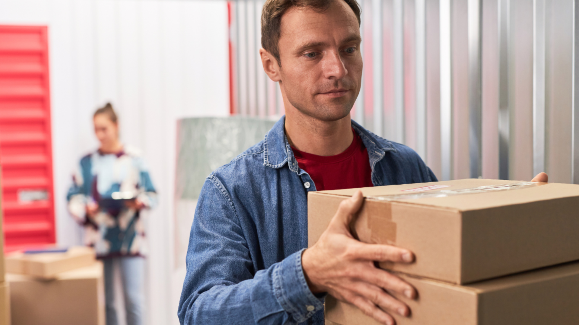 A person in a denim shirt carries a stack of cardboard boxes in a storage facility with another person blurred behind them.
