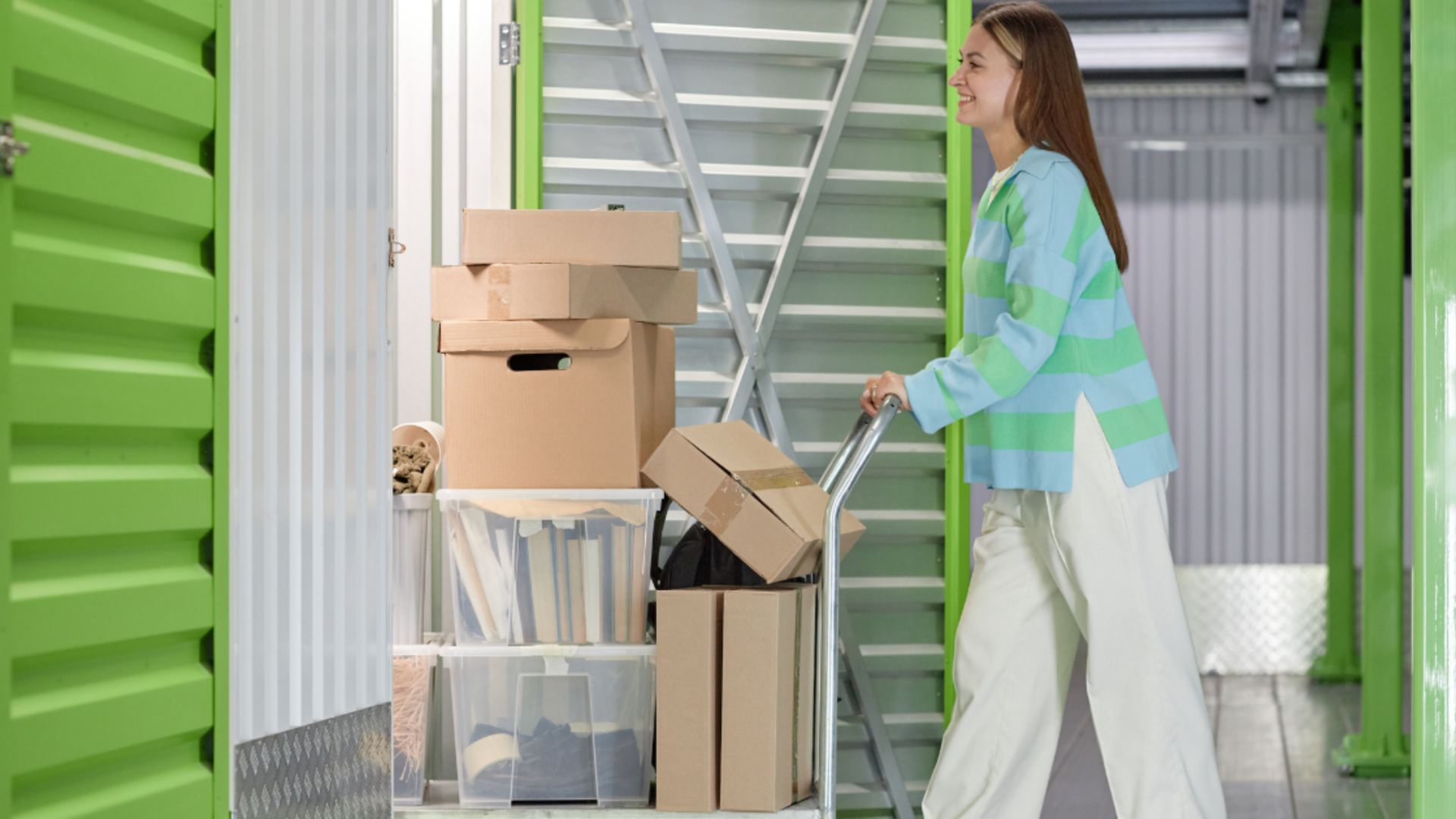 A person wearing a striped sweater pushes a cart loaded with cardboard boxes and storage bins into a storage facility.